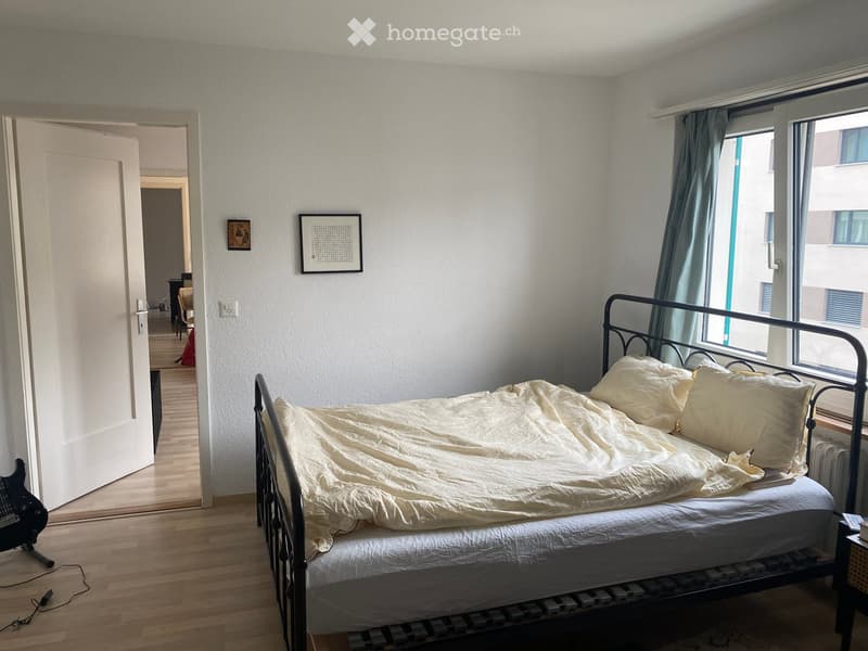 Bedroom with a single bed, nightstand, guitar, and picture frames on the wall. The room has wooden flooring and large windows with curtains.