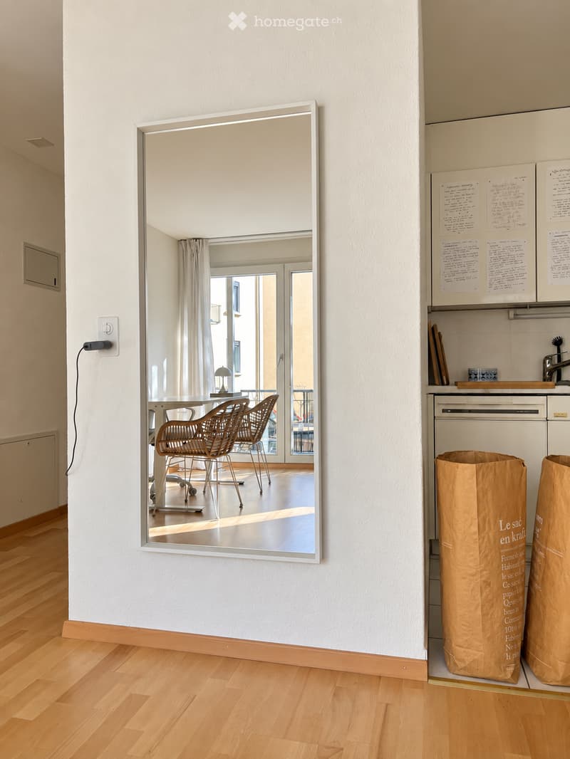 A hallway with light wooden flooring, white walls, and a large mirror reflecting a dining area with wicker chairs and a table. There are two large brown paper bags on the floor and a kitchen area visible to the right.