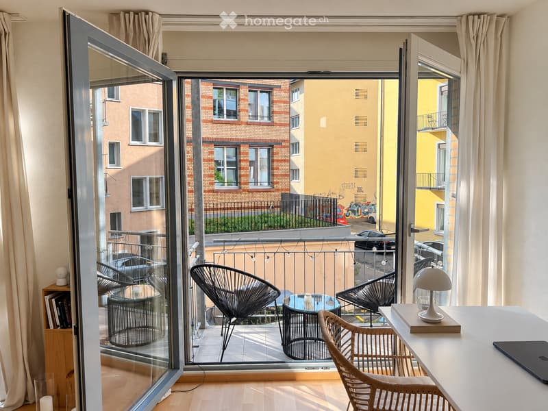 Modern room with large balcony access, featuring a white desk with a laptop, a lamp, and a bookshelf. The balcony has a glass table and several chairs.