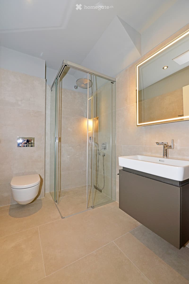 Modern bathroom with beige tiles, glass-enclosed shower, toilet, sink with modern faucet, and illuminated mirror.