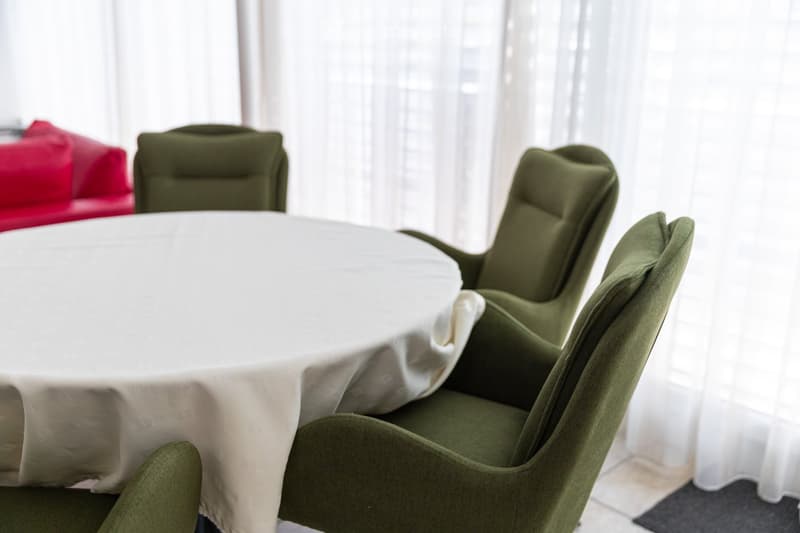 A dining area with a round table covered in a white tablecloth and several green upholstered chairs arranged around it. The background shows a red couch and large windows with white curtains.