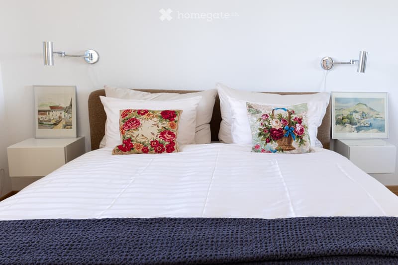 A neatly made bed with white linens, decorative pillows, and a blue throw blanket. Two nightstands with picture frames and wall-mounted lamps flank the bed.