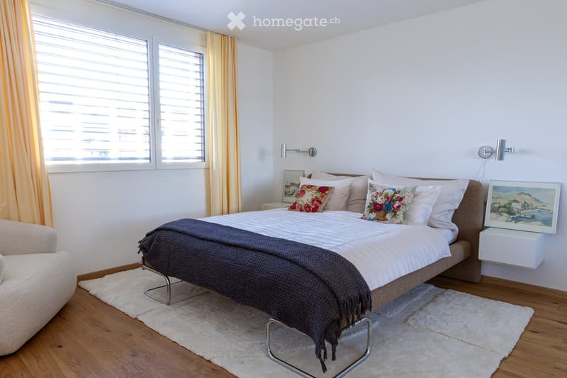 Modern bedroom with a large bed, decorative pillows, a dark throw, a white armchair, nightstand, and artwork on the wall. The room features wooden flooring and large windows with blinds.