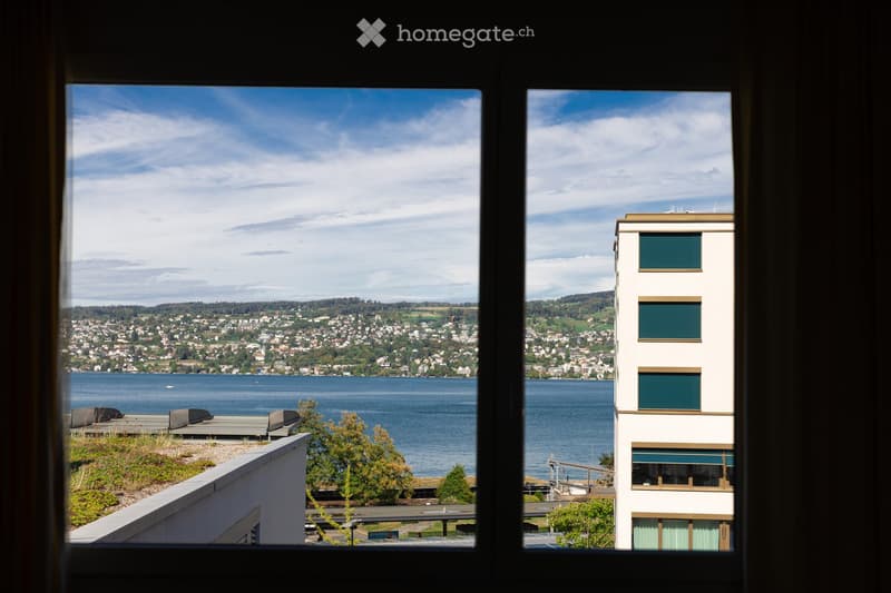 View from a window showing a cityscape across a body of water, with a modern building in the foreground and a distant landscape.