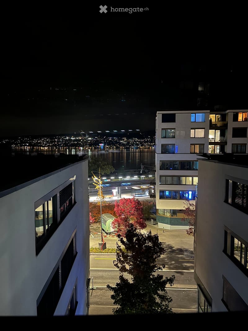 Nighttime view from a balcony showing a cityscape with illuminated buildings, a waterfront, and a train station in the distance.