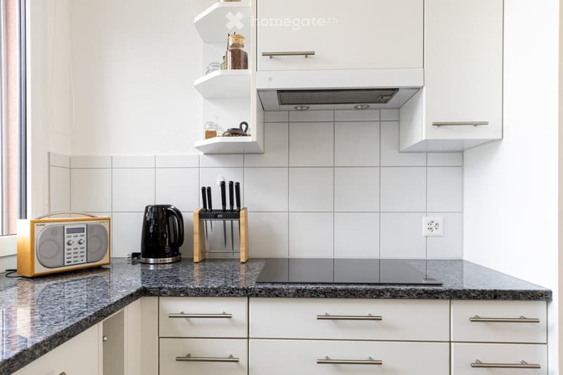 Modern kitchen with white cabinets, granite countertop, built-in range hood, and various appliances including a kettle and radio.