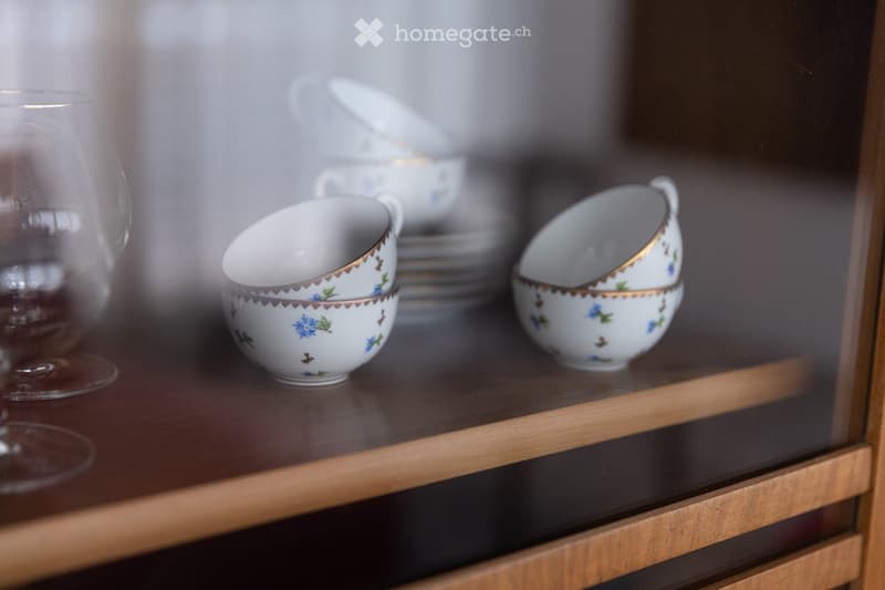 A wooden cabinet displays decorative cups and plates with floral patterns, along with a clear glass on the left side.