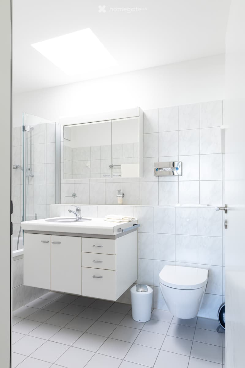 Modern bathroom with white tiles, a white sink with a cabinet, a toilet, a mirror, and a glass shower enclosure.