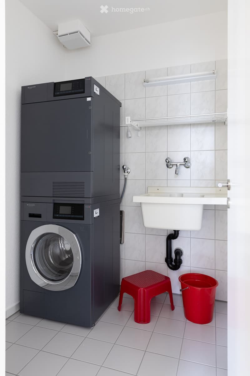 A modern bathroom with a washing machine, a sink, a red stool, and a red bucket. The walls are tiled, and there is a white shelf above the sink.