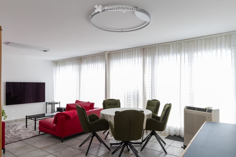 Modern living room with a red sofa, a round dining table with green chairs, a television mounted on the wall, large windows with white curtains, and an air conditioner unit.