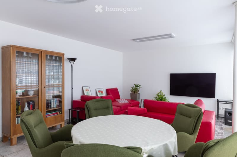 Modern living room with white walls, a red sofa, green armchairs, a round table with a white tablecloth, a wooden cabinet, a television mounted on the wall, and a floor lamp.