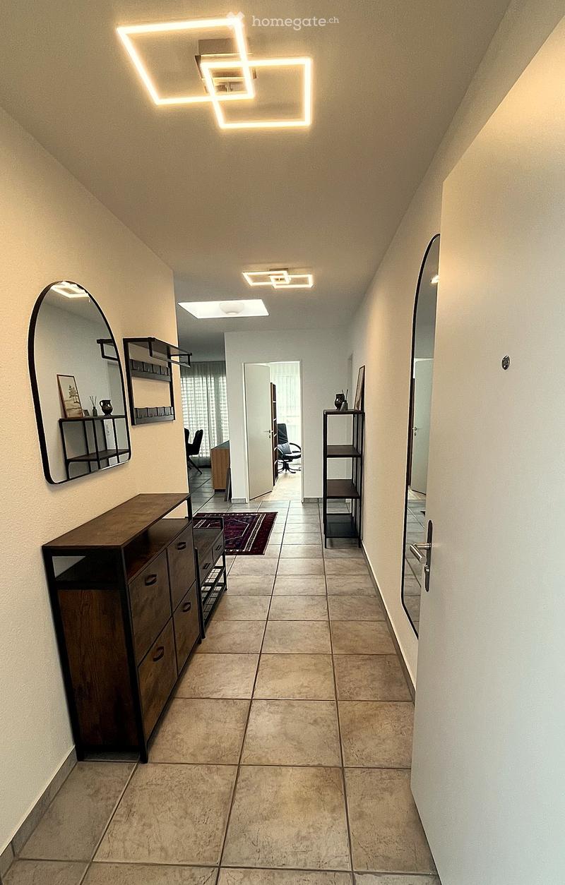 Modern hallway with beige tiled flooring, white walls, and geometric LED ceiling lights. Features a wooden cabinet, a small shelf, and a mirror on the left, with a doorway leading to another room.