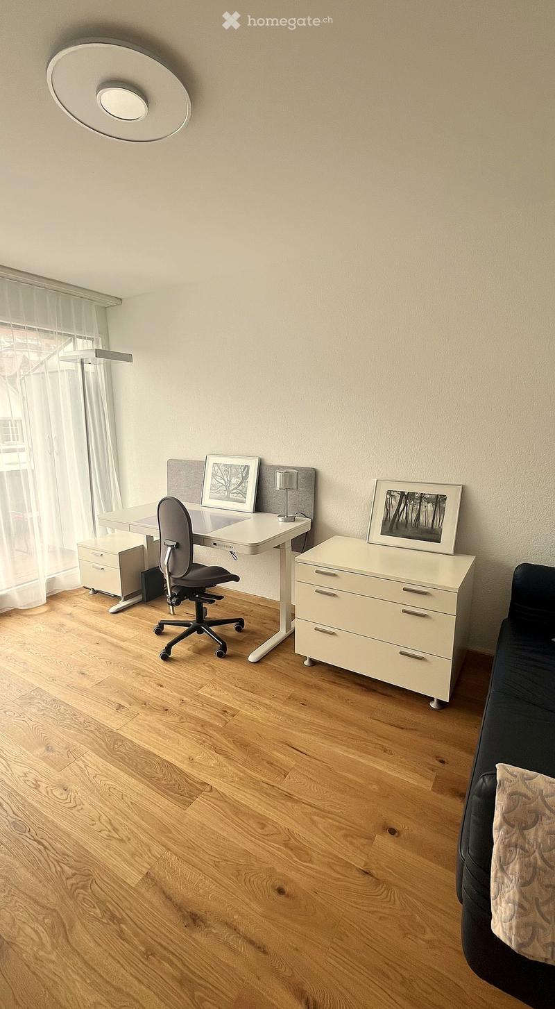 A modern, well-lit room with wooden flooring, a white desk, a black office chair, a white cabinet, a sofa, and framed artwork on the walls.