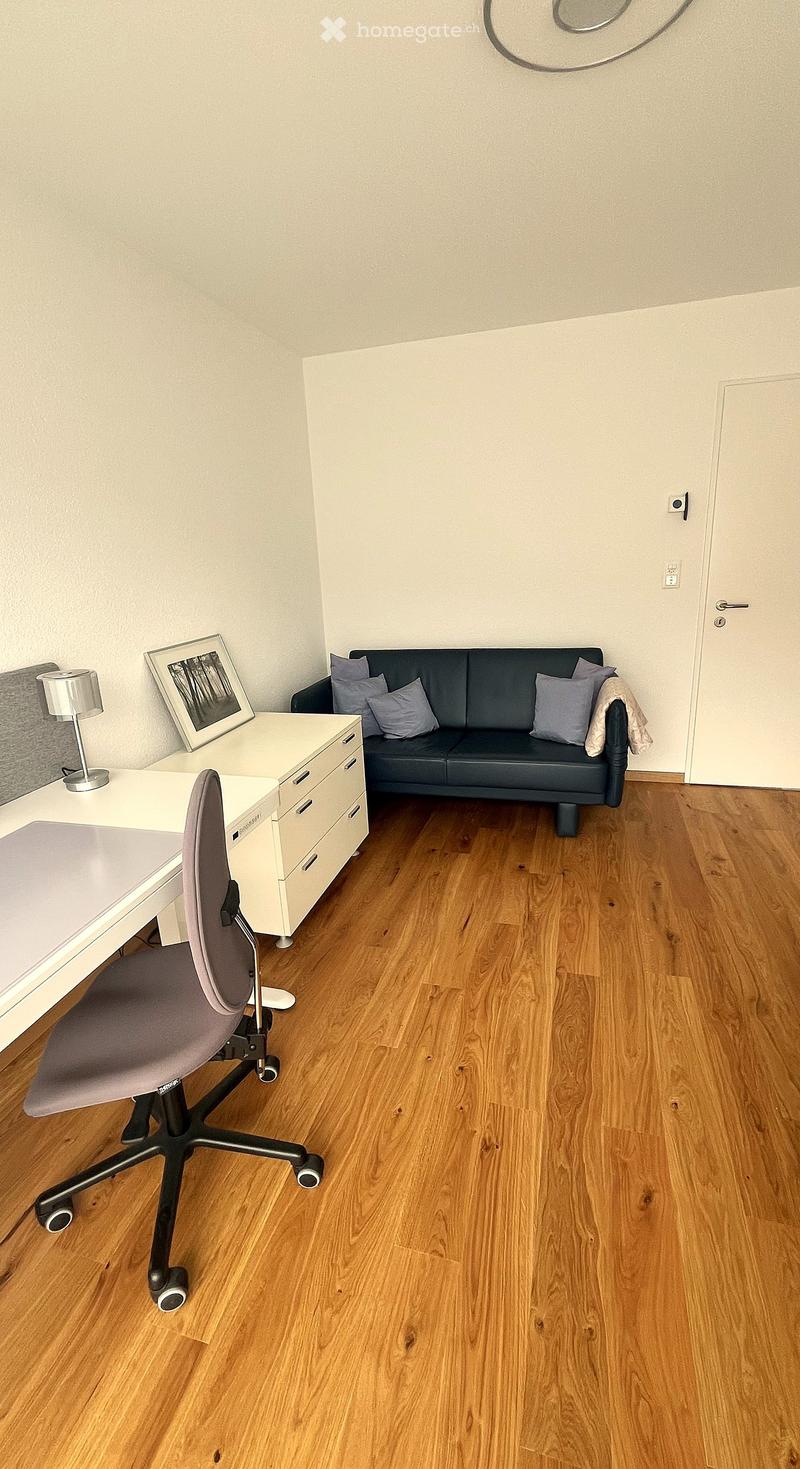 A modern, well-lit room with wooden flooring, a grey sofa with cushions, a white desk with a chair, a lamp, and a picture frame on the wall.