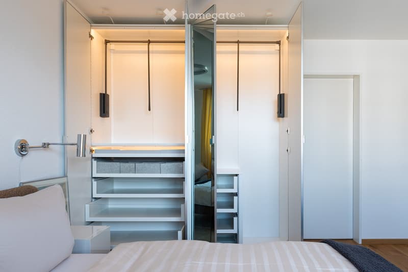Modern bedroom with a neatly made bed, white nightstand, built-in wardrobe with mirrored doors, and a sleek wall-mounted lamp.