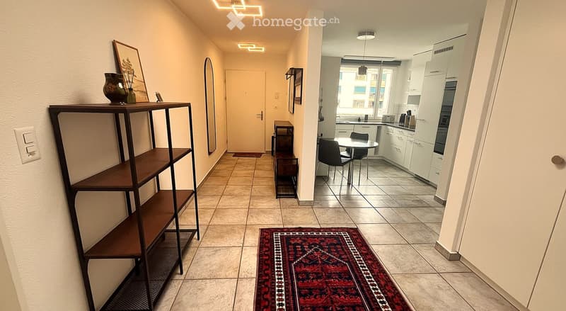 Hallway with tiled floor, wooden shelving unit, small table, and a red patterned rug leading to a kitchen area with white cabinets, a dining table, and chairs.