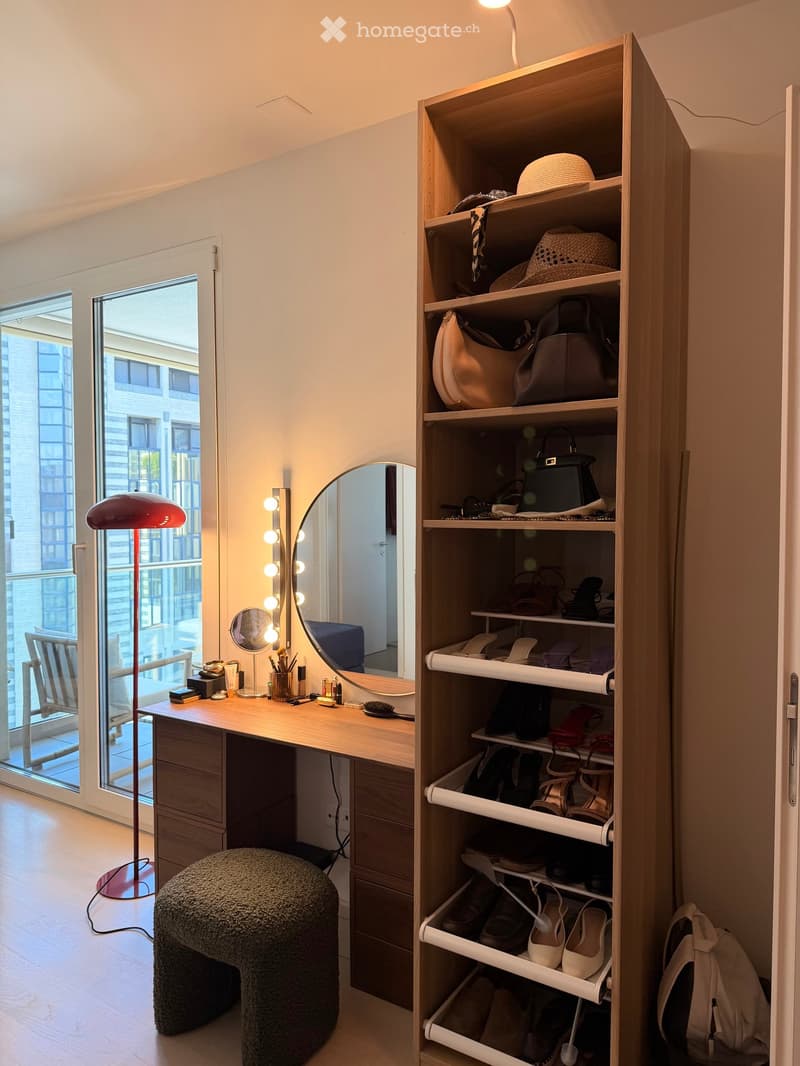 A well-organized dressing area with a vanity table, mirror, floor lamp, and a tall wardrobe filled with shoes, handbags, and hats. A round, fluffy stool is placed in front of the vanity.