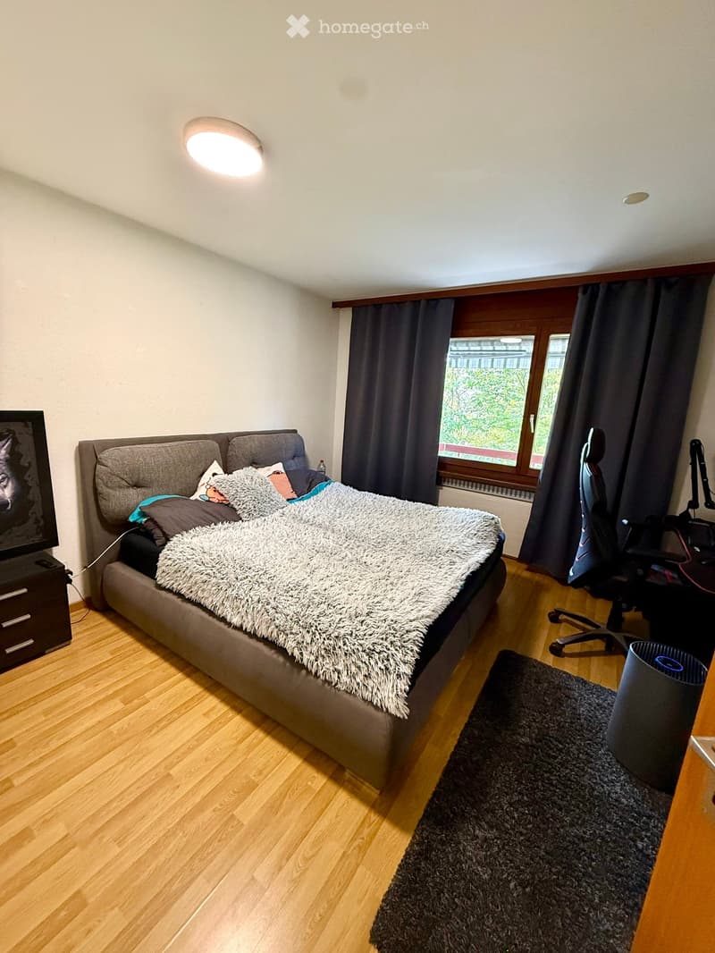 A modern bedroom with a large bed, fluffy white throw, pillows, a TV screen, a chair, a small table, and a window with dark curtains.