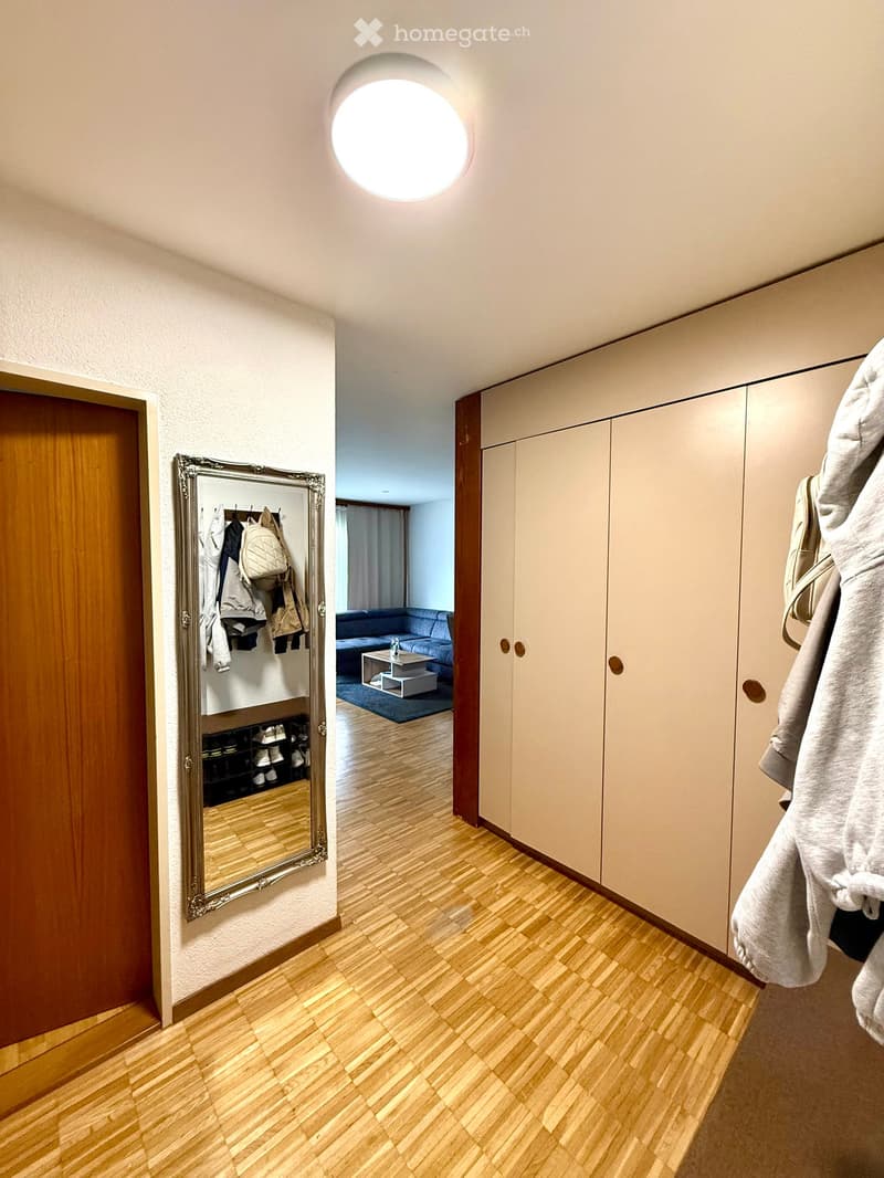 A well-lit hallway with wooden flooring, white walls, and built-in storage units. A large mirror with a decorative frame is mounted on the wall, and a glimpse of a living room area with a sofa and coffee table is visible through an open doorway.