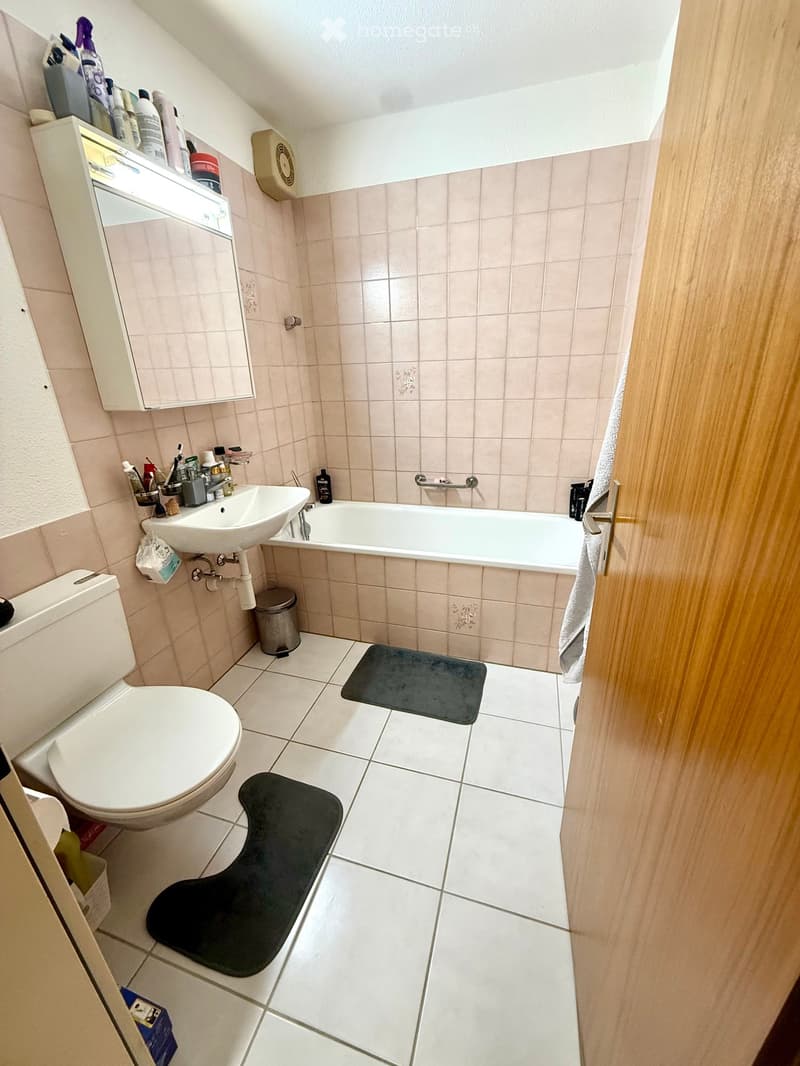 Bathroom with a toilet, bathtub, sink, mirror, and a small cabinet. The floor is tiled, and there are two black bathmats placed in front of the toilet and bathtub.