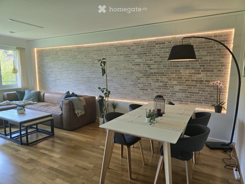 Modern living and dining area with a brick accent wall, wooden flooring, a brown leather sofa, a white dining table with chairs, and decorative plants.