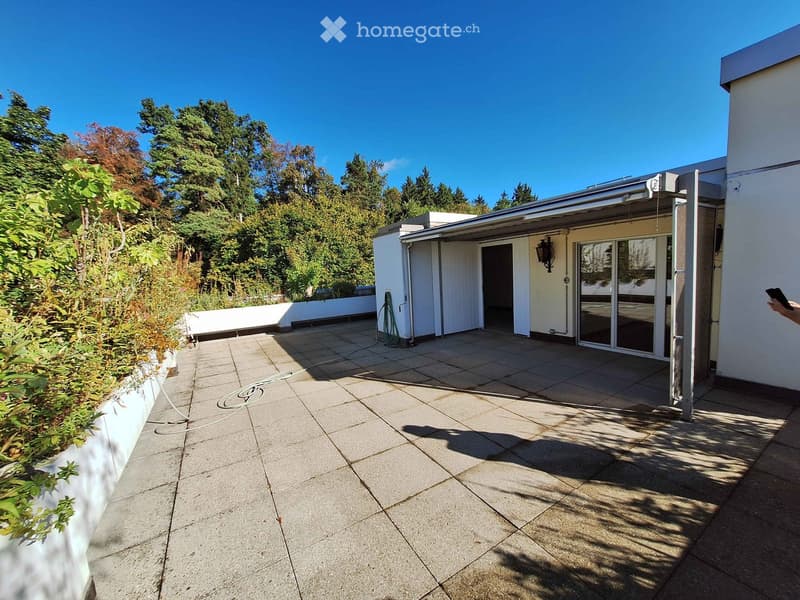 A modern outdoor patio with tiled flooring, a covered entrance, and surrounding greenery.