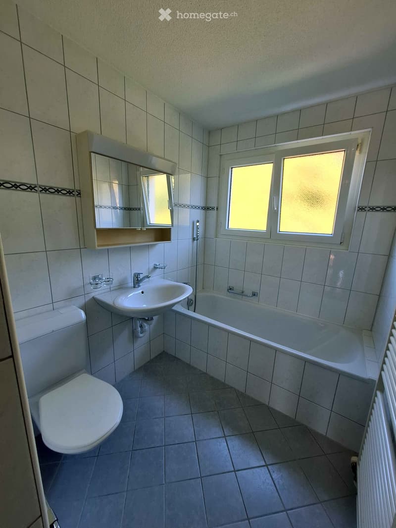 Bathroom with a toilet, sink, bathtub, mirror, and a window with frosted glass. The walls and floor are tiled, and there is a radiator on the right side.