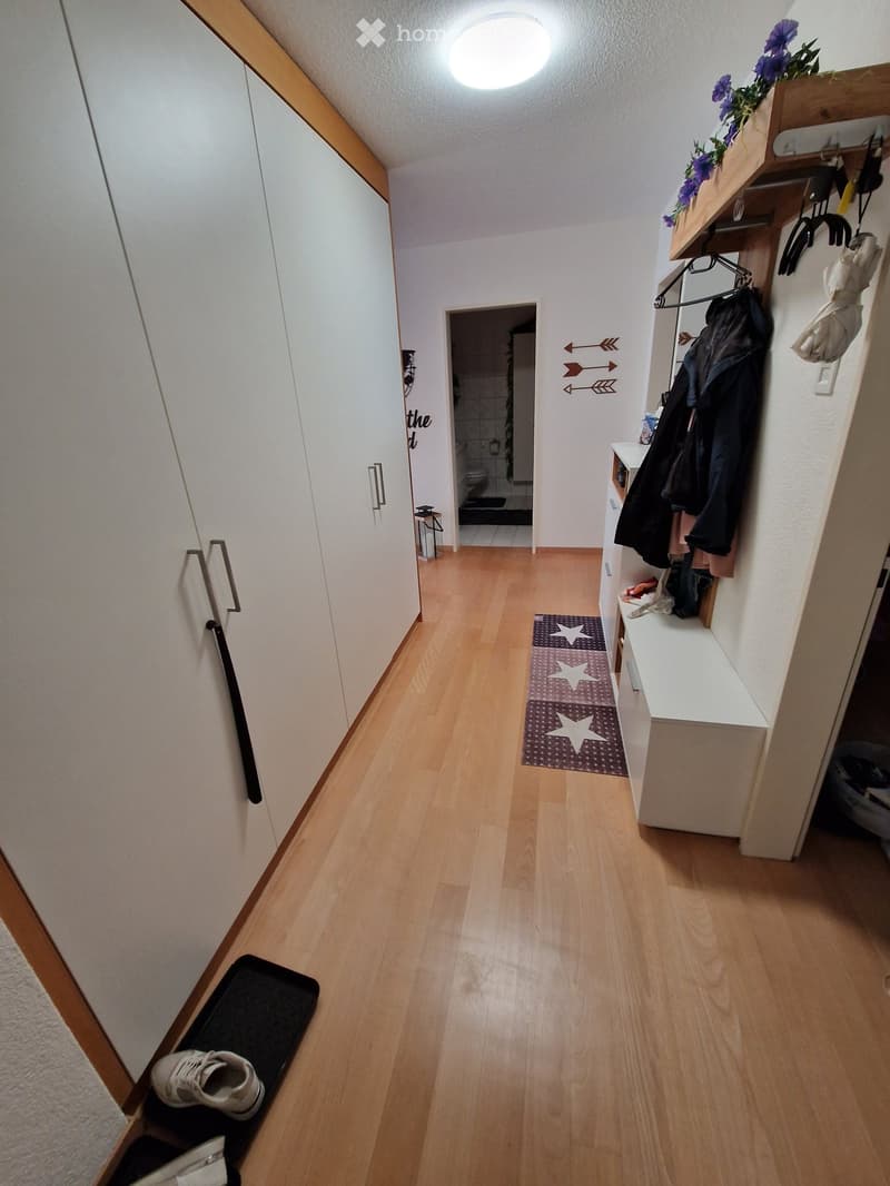 Clean hallway with light wooden flooring, white walls, coat rack with jackets, small shelf with decorative items, and a glimpse of a bathroom at the end.