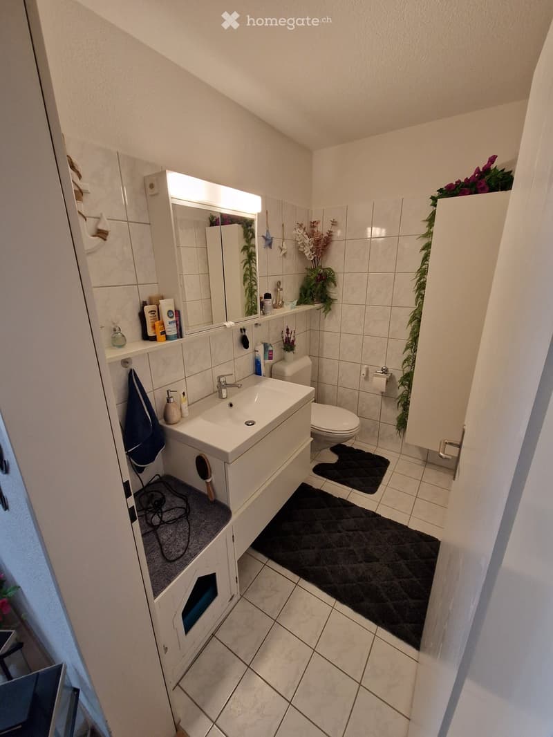 Modern bathroom with white tiles, a sink, toilet, mirror, and a small cabinet. There are two black rugs on the floor and a towel hanging on the side.