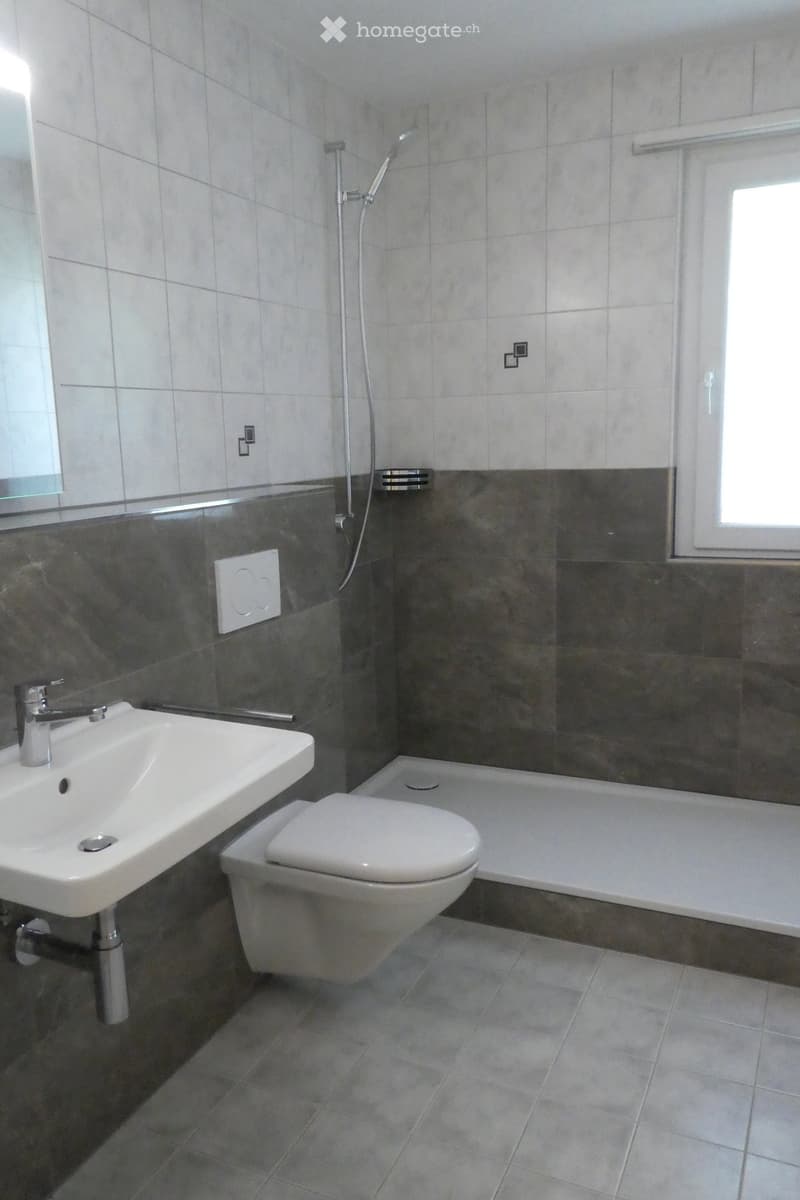 Modern bathroom with a white sink, toilet, bathtub, and showerhead. Grey tiled walls and floor, with a window providing natural light.