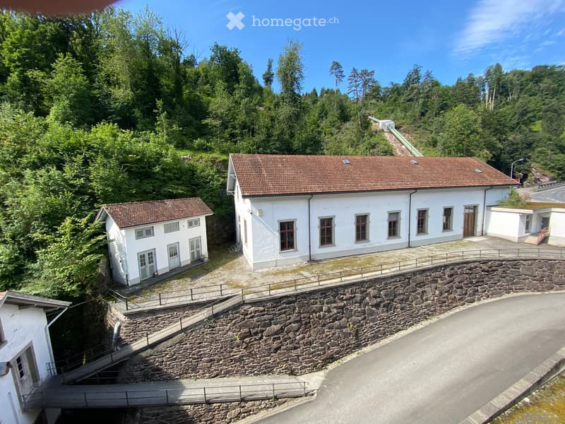 Aerial view of a white building with a brown tiled roof, surrounded by greenery and a stone retaining wall.