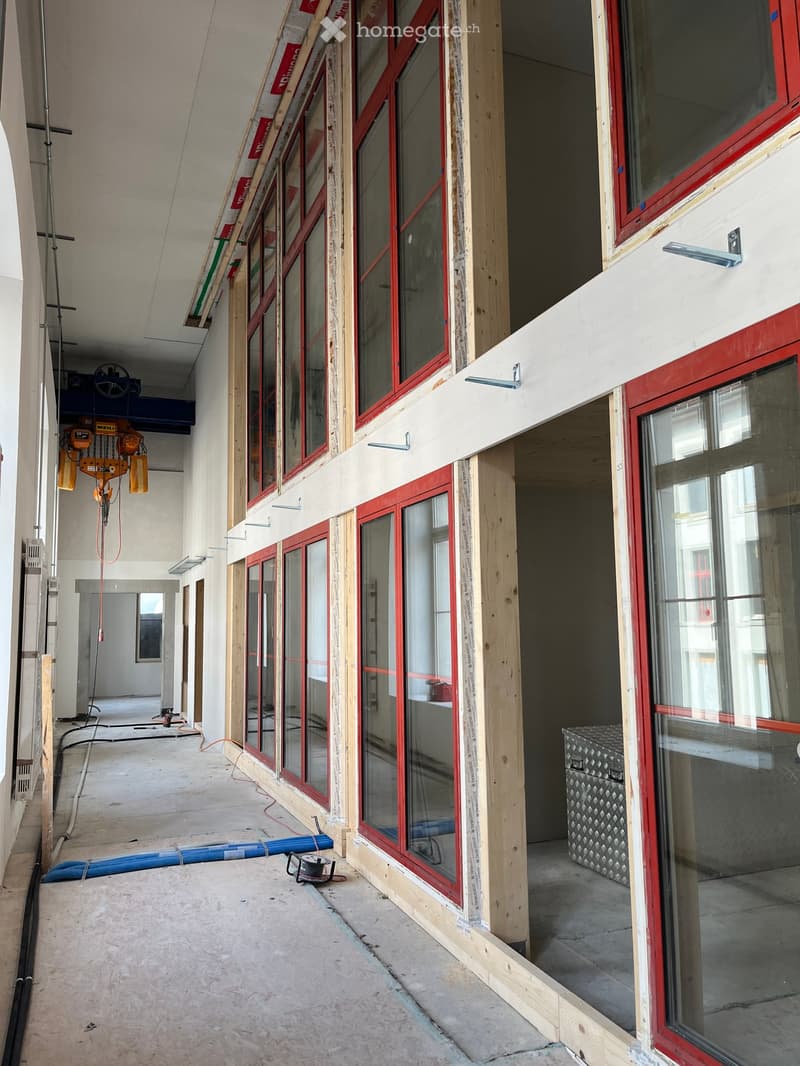 Empty corridor with exposed wooden framing and large windows with red frames, construction materials and tools visible.