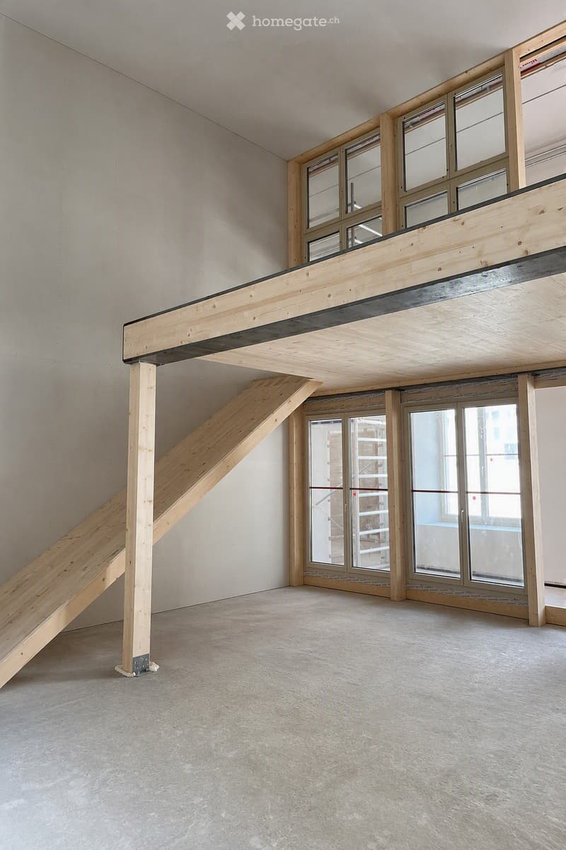 Empty room with exposed wooden beams, a staircase leading to an upper level, and large windows with glass panels.