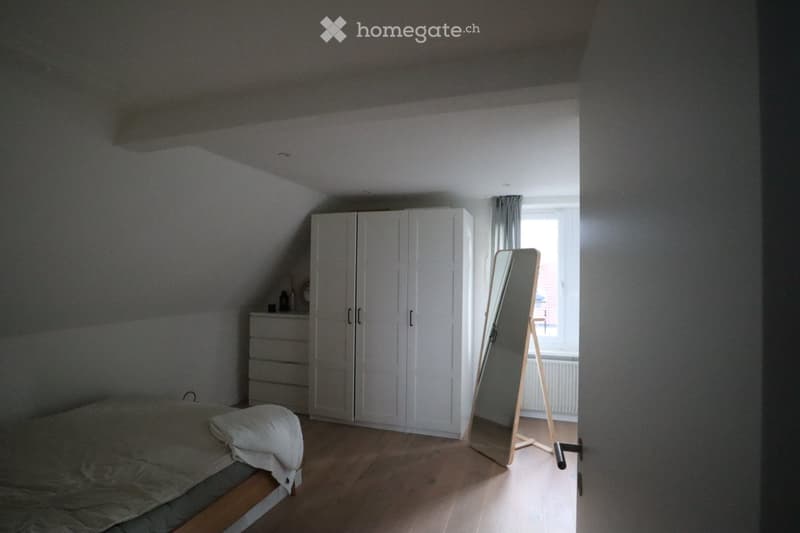Bright bedroom with wooden flooring, a large bed with white bedding, a full wardrobe, a dresser, and a standing mirror near the window.