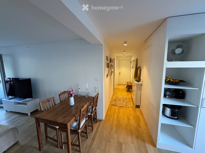 A well-lit, modern hallway leading to a dining area with a wooden table, chairs, and a vase of flowers. A white cabinet with a fan and kitchen appliances is visible, along with a television on a stand and a small bench in the hallway.