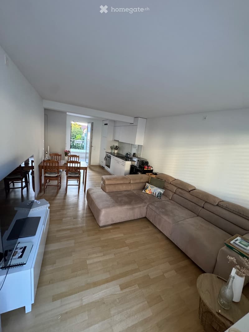 A modern living room with a large sectional sofa, coffee table, side table with vase, dining area with chairs and table, and an open kitchen area with cabinets, oven, microwave, and refrigerator.