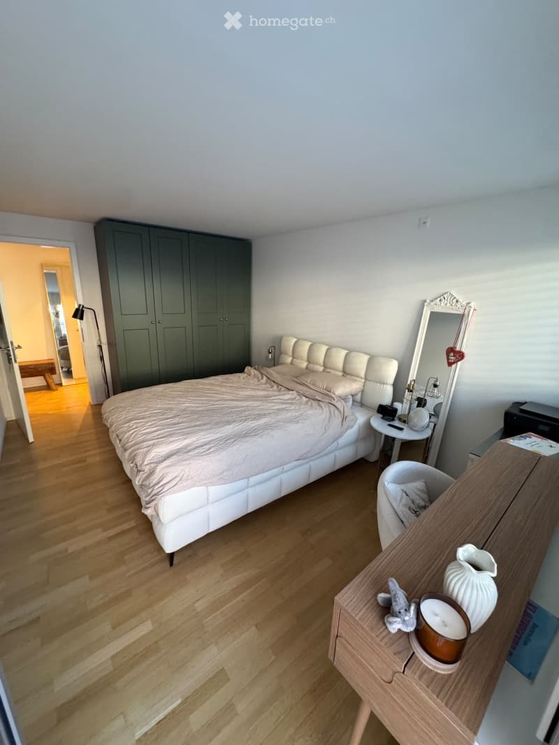 Modern bedroom with a white upholstered bed, wooden floor, large mirror, side table, and a small chair. A cabinet and a lamp are also visible.