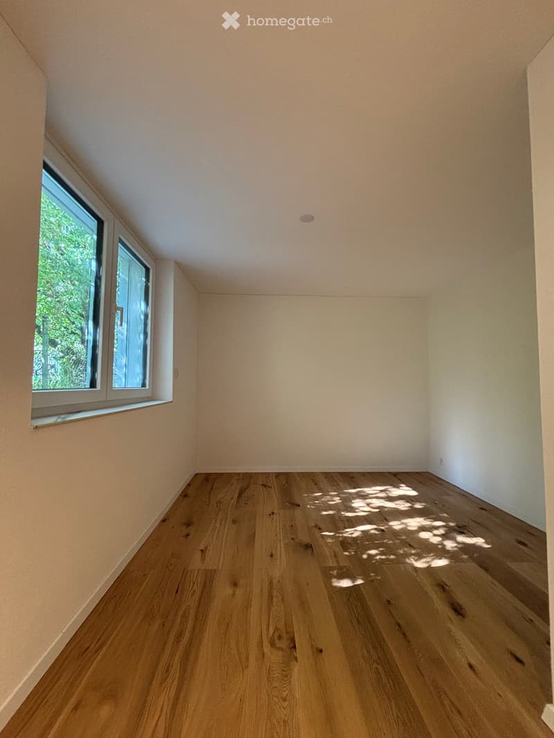Empty room with wooden flooring and light-colored walls, featuring a large window with a view of greenery outside.