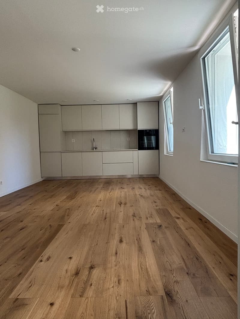 Empty kitchen with light wooden flooring, white cabinets, and a window on the right side.