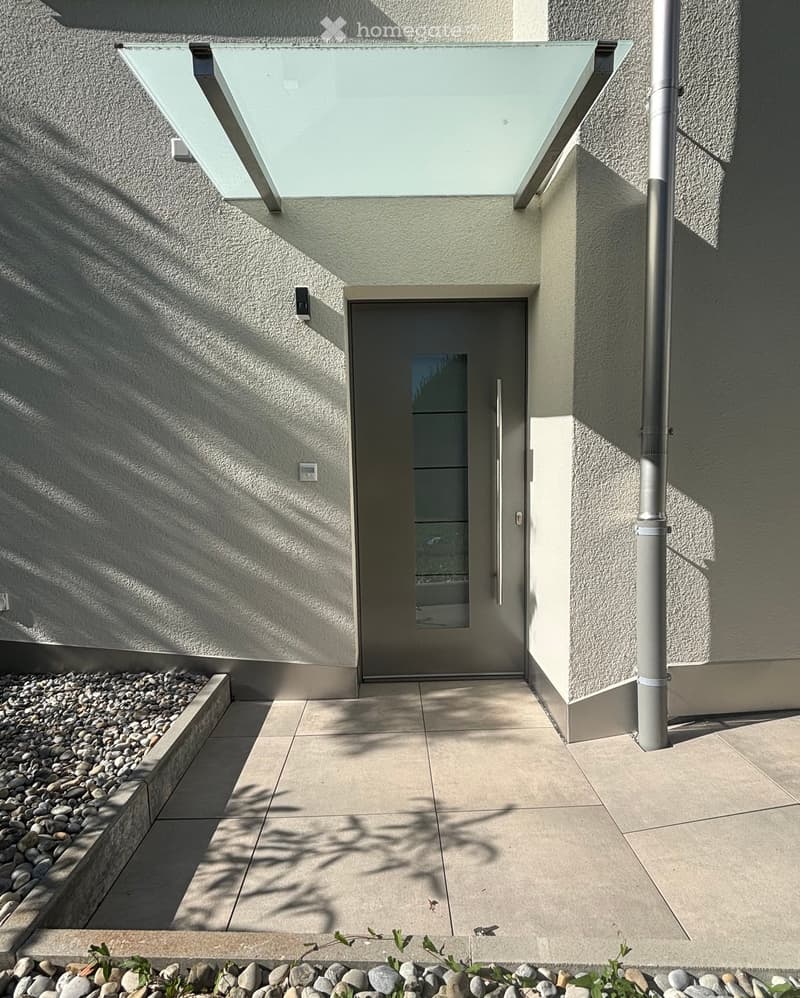 Modern building entrance with a sleek glass door, textured light gray walls, and a raised ground floor with a small garden area featuring rocks and sparse greenery.