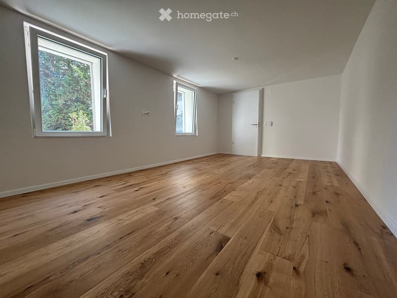 Empty room with wooden flooring, white walls, and large windows with a view of greenery outside.
