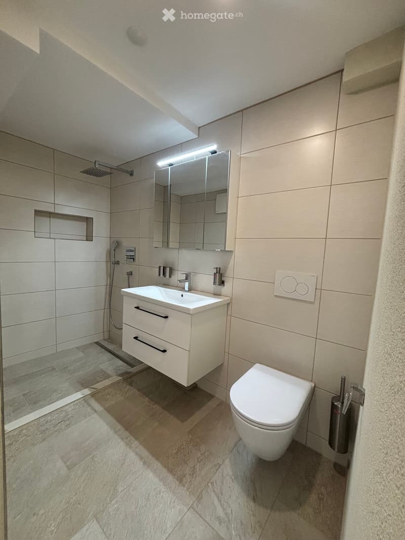 Modern bathroom with a white sink, toilet, mirror, and shower area. Features light-colored tiles and a sleek design.