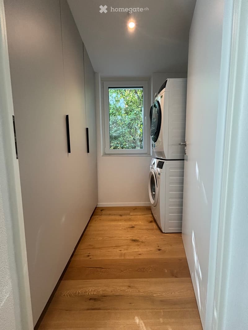 Narrow hallway with wooden flooring, white walls, and a window showing greenery. A private washing machine is placed against the wall.