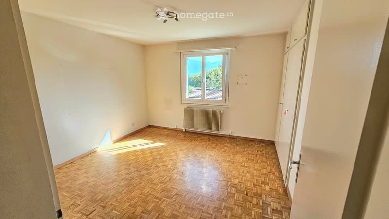 Empty room with wooden parquet flooring, white walls, a window with a view, and a radiator below the window.