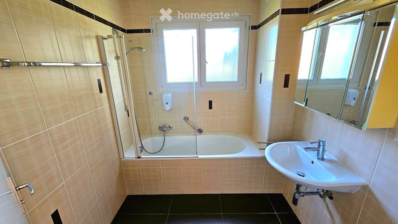 Modern bathroom with a bathtub, sink, mirror, and shower area. Features beige tiled walls, black tiled floor, and a window above the bathtub.