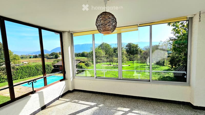 Bright, unfurnished room with large windows offering a scenic view of a green landscape and mountains. A swimming pool is visible outside.