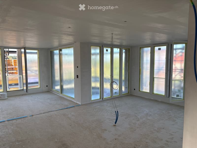 Empty room with bare walls, concrete floor, and large windows with frosted glass. Exposed electrical wiring and construction materials are visible.