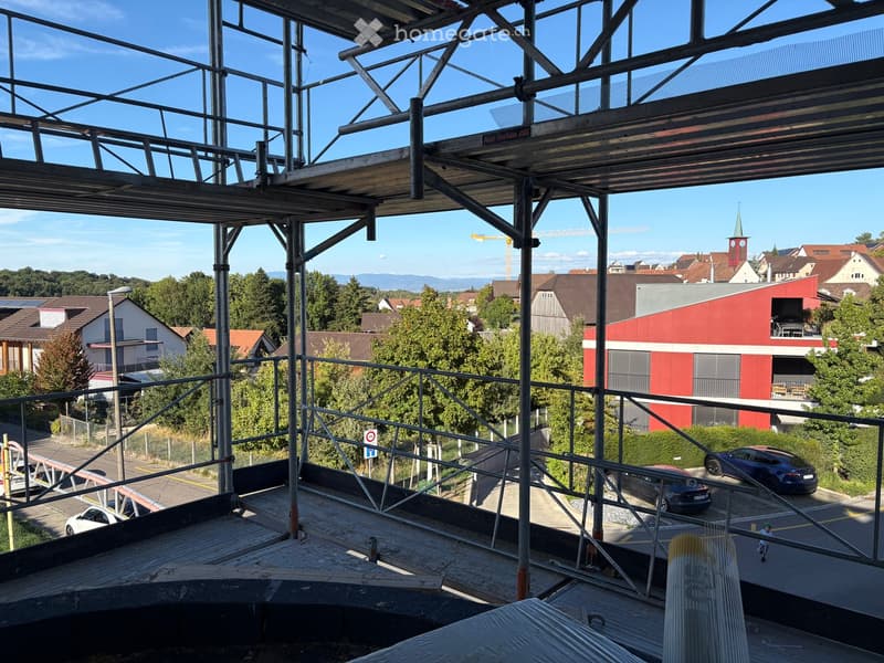 View from a rooftop showing a construction site with scaffolding, metal beams, and a partially built structure. The background includes a town with houses, trees, and a church tower.