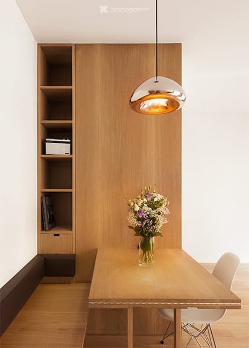 Modern dining area with a wooden table, white chair, built-in wooden bookshelf, and a hanging pendant light.