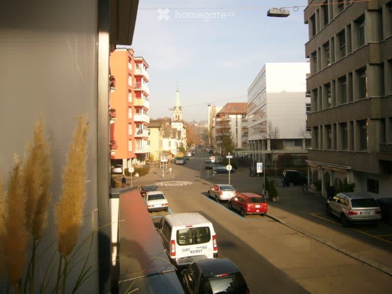 Balcony overlooking a city street with parked cars, buildings, and a distant church spire.
